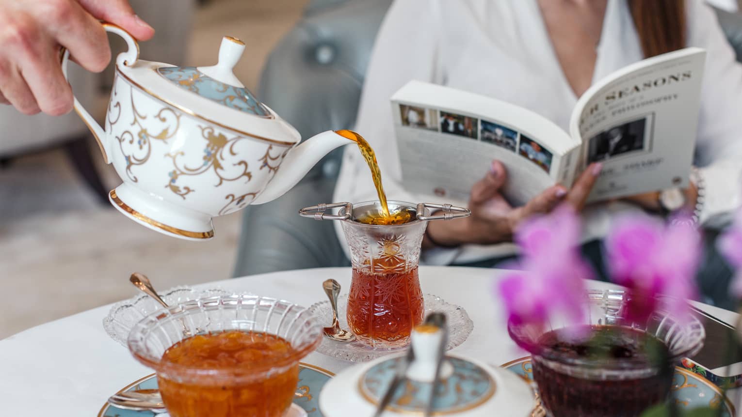 Woman reads book as server pours afternoon tea from gold and white pot