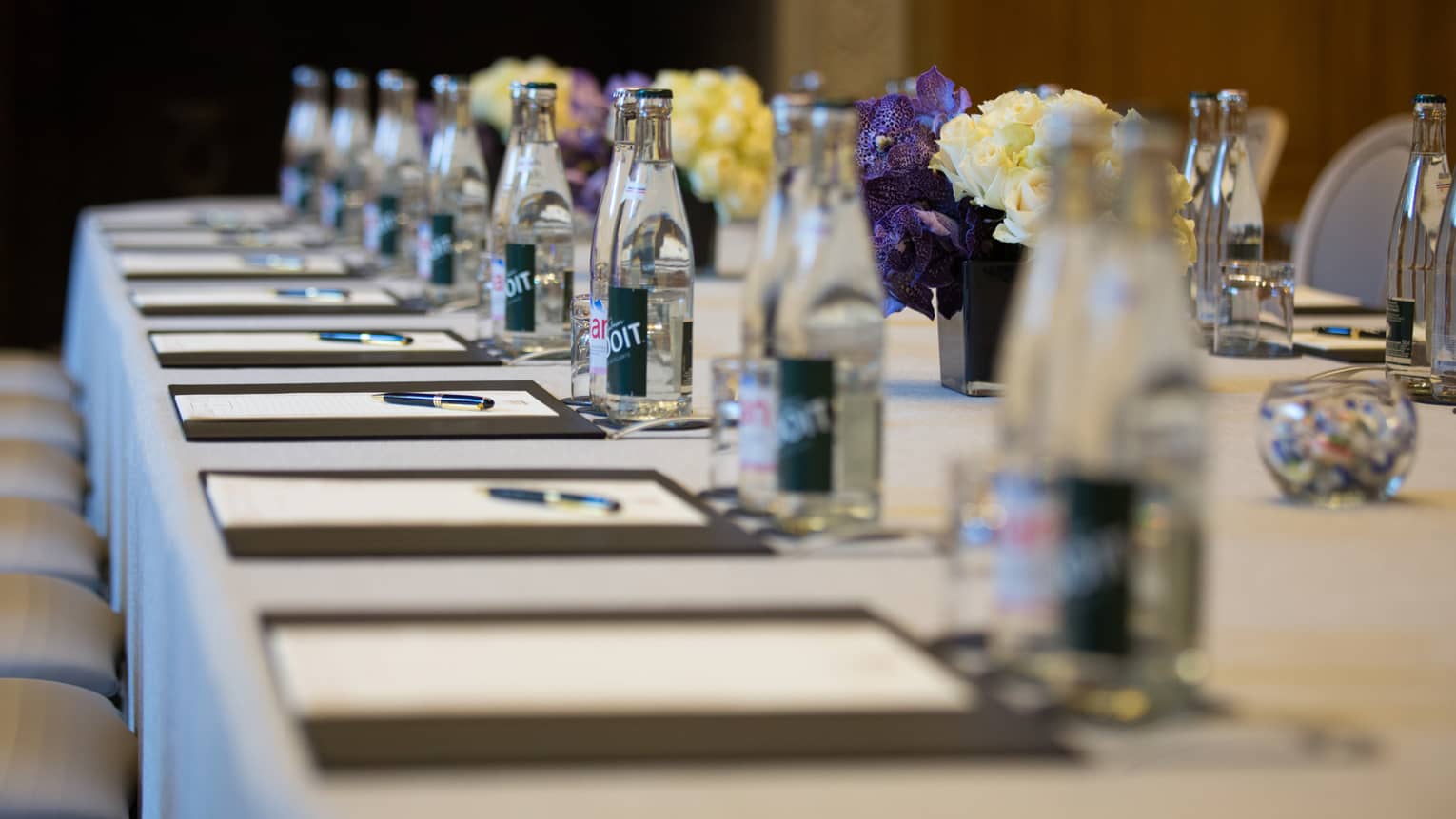 Row of meeting agendas, glass bottles, small vases with flowers on boardroom table