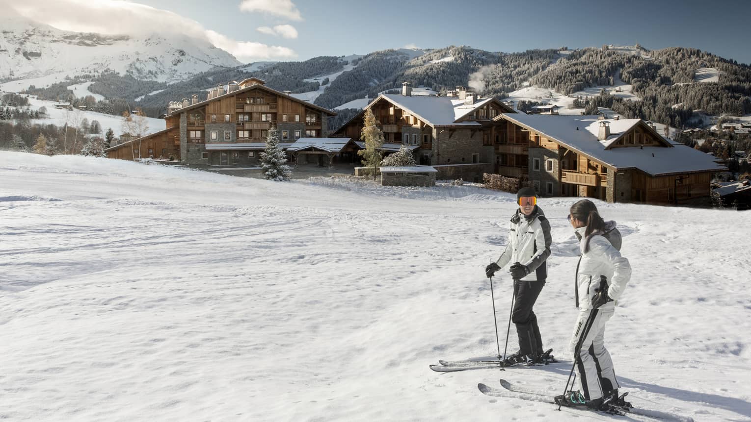 Man, woman on skis at top of snowy hill, Four Seasons Hotel Megeve in background