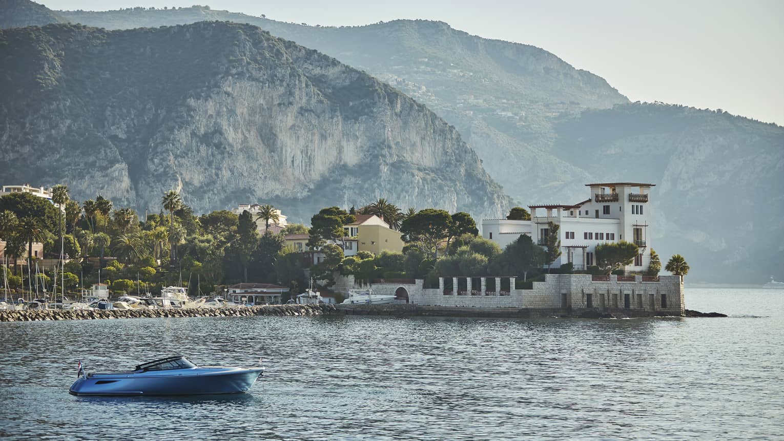 Cliffs and hills loom above a forested village along the water; a speed boat is anchored in front of a stone breakwater.