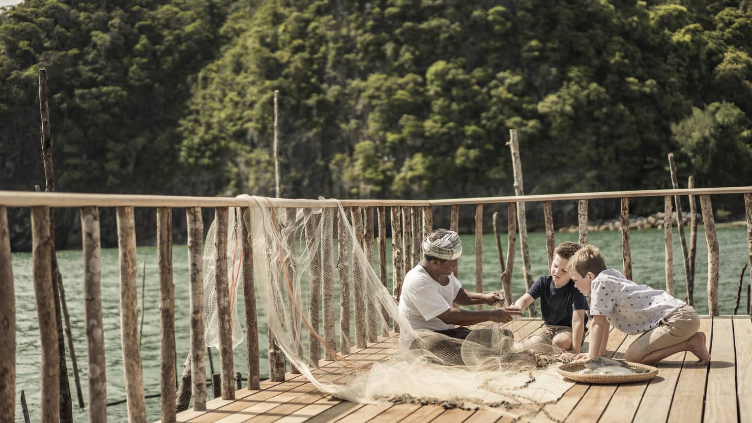 A fisherman and two boys on a dock looking at the fish they caught in their net at Four Seasons Langkawi