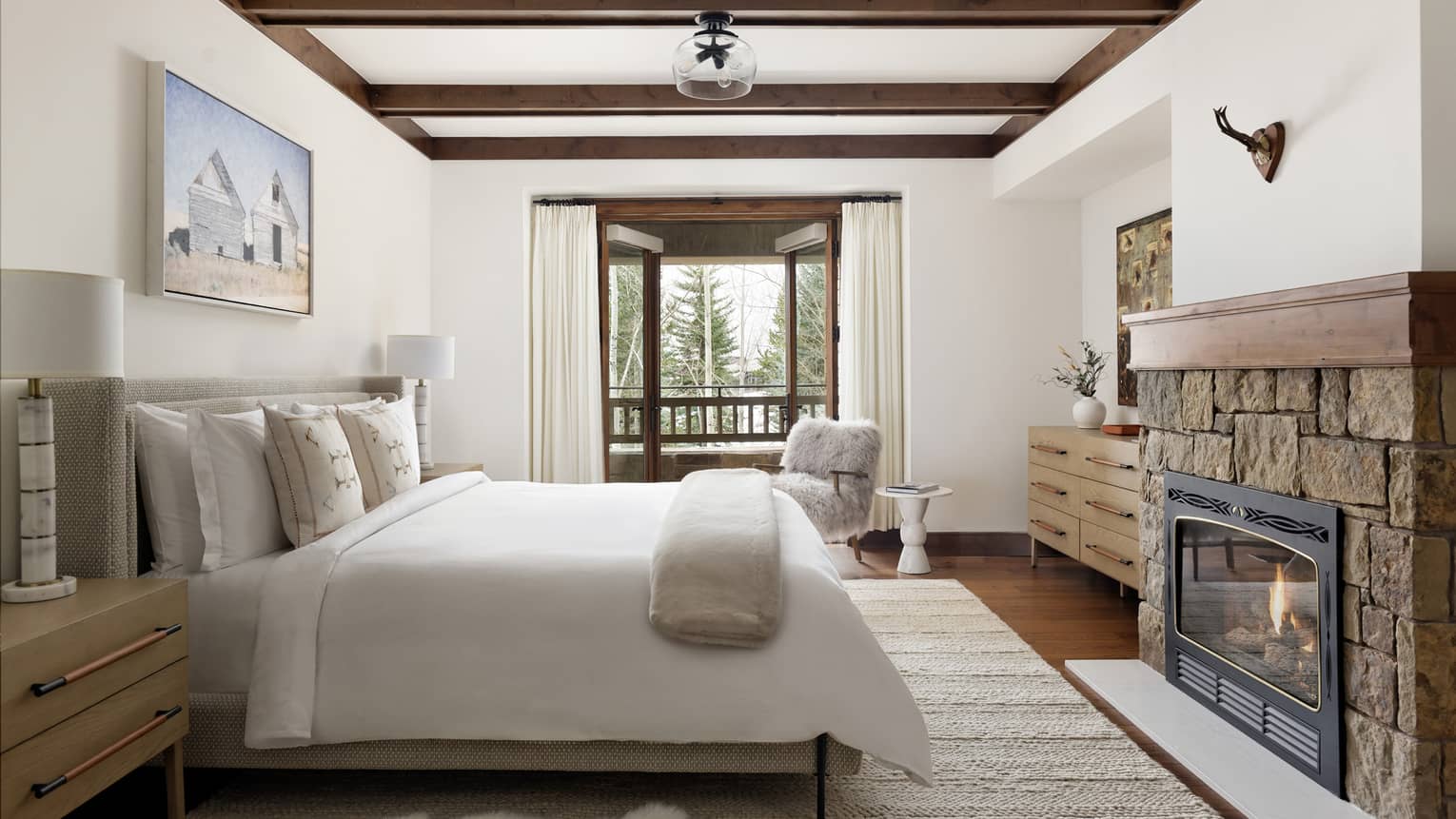 Bedroom with a king bed, nightstands with lamps, wall artwork, stone fireplace, dresser, fluffy armchair, and balcony 