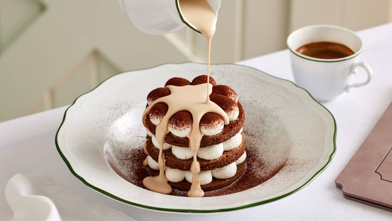 Dessert with alternating layers of dollopw of cream and chocolate cake sit in a white bowl as cream is being poured over it