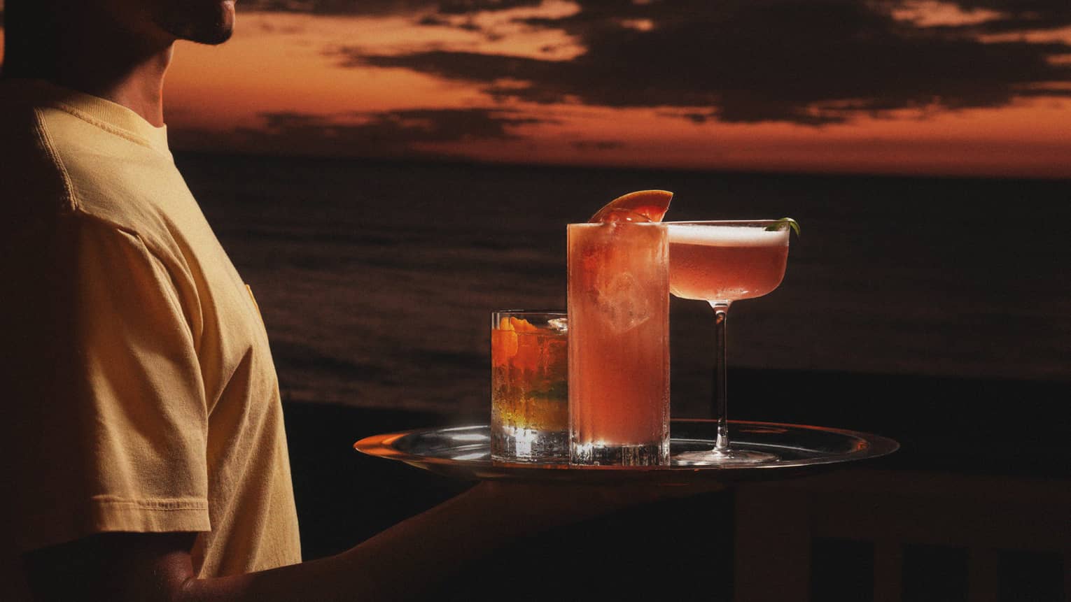 Server carries three cocktails on a tray with an orange-coloured sky in the background