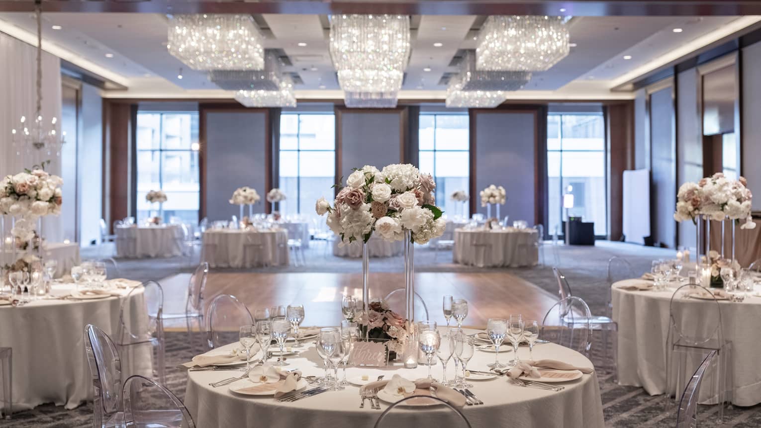 A well lit ballroom with ground tables and flower centerpieces and a section of hardwood floor in the middle.