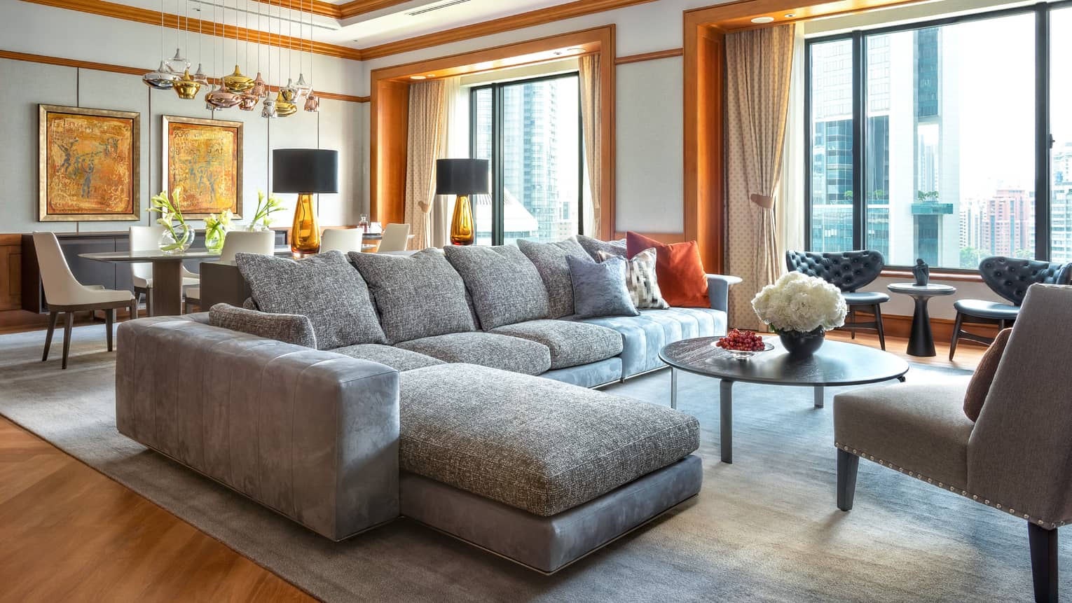 A modern living room with a large grey sectional sofa, accented with colourful pillows, a round coffee table and floor-to-ceiling windows. A dining area with decorative lighting and artwork is visible in the background.