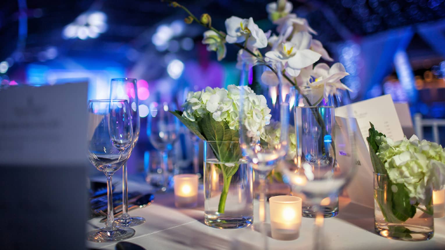 Close up of white flowers, glassware on wedding reception dining table, blue lights in background