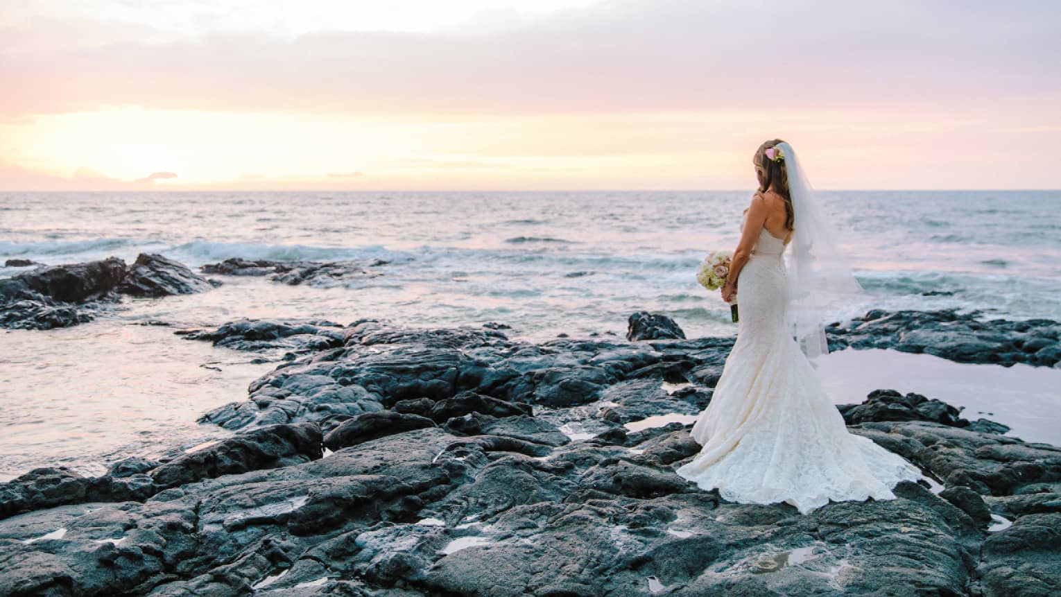 Bride in white wedding gown poses on black lava rocks by ocean