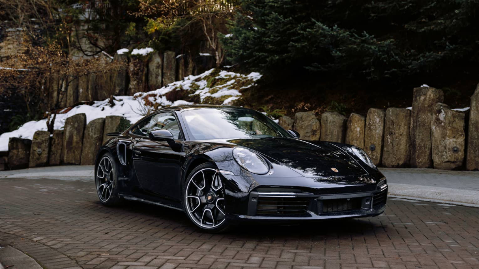Black Porsche coupe sits in brick driveway, snow on the ground behind it
