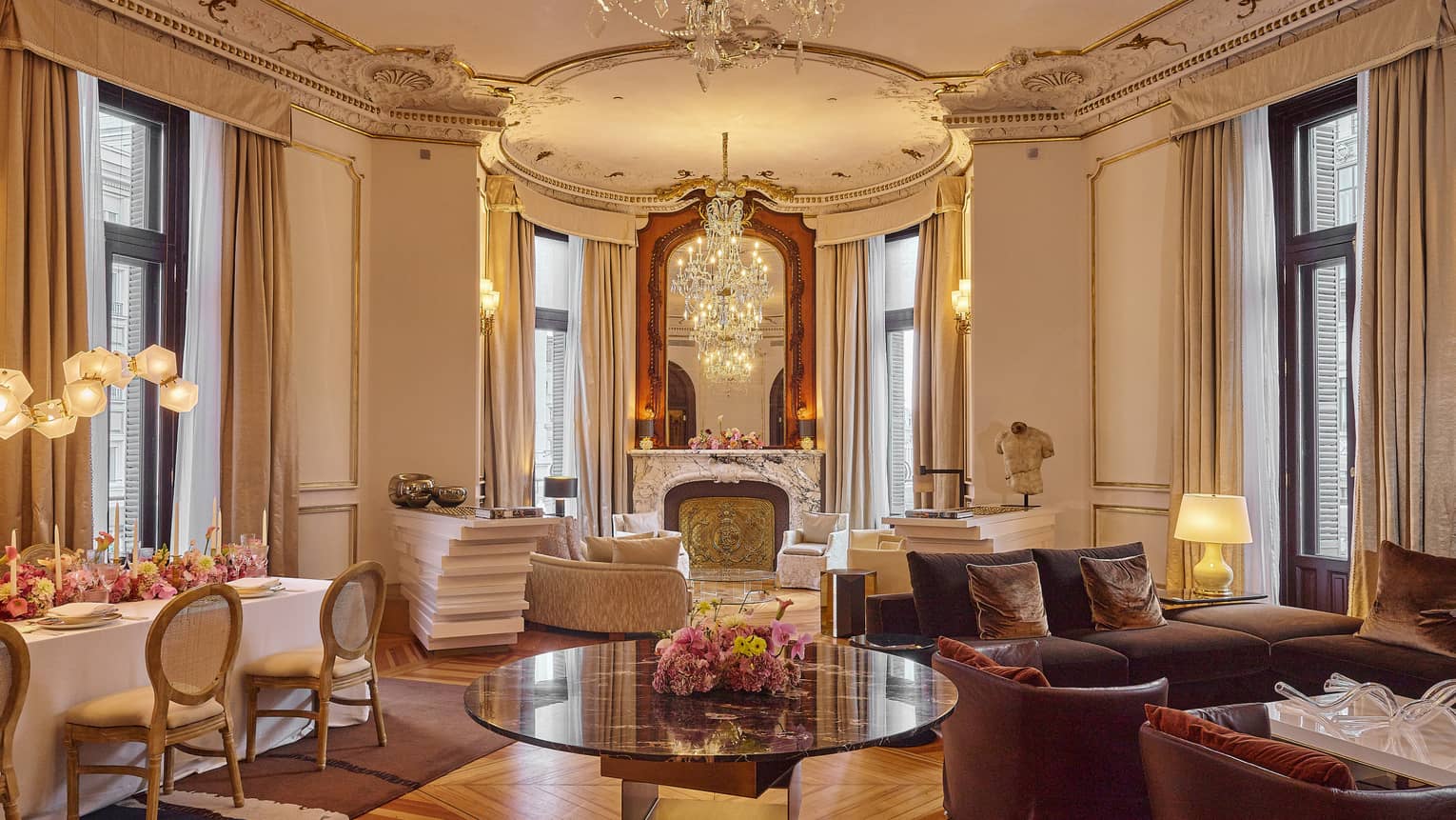 Large Regency-style living and dining area with tall ceilings, ornate finishes, two seating areas, a rectangular dining table and four sets of black-framed, floor-to-ceiling windows with beige curtains pulled to the side