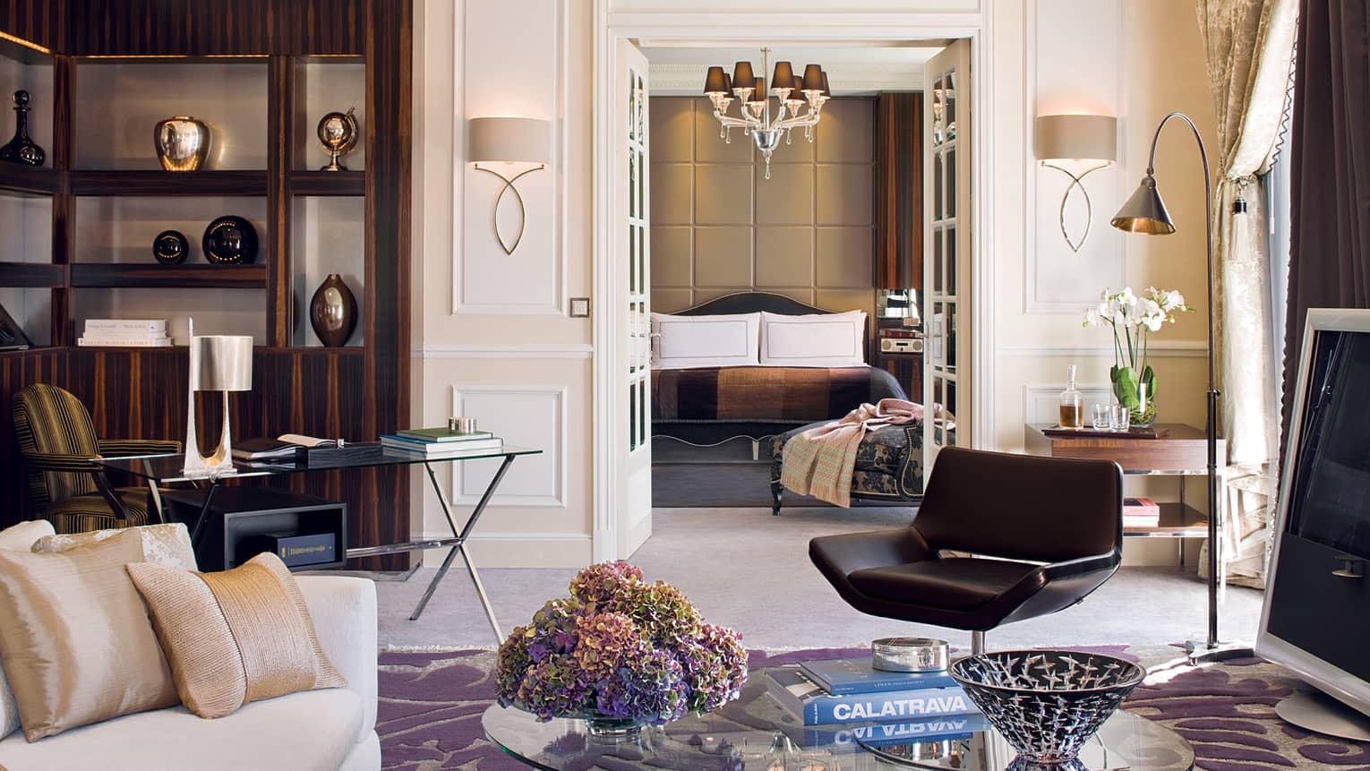 Hotel living room with coffee table, book, retro black chair, glass desk in front of shelf, vases, bedroom door