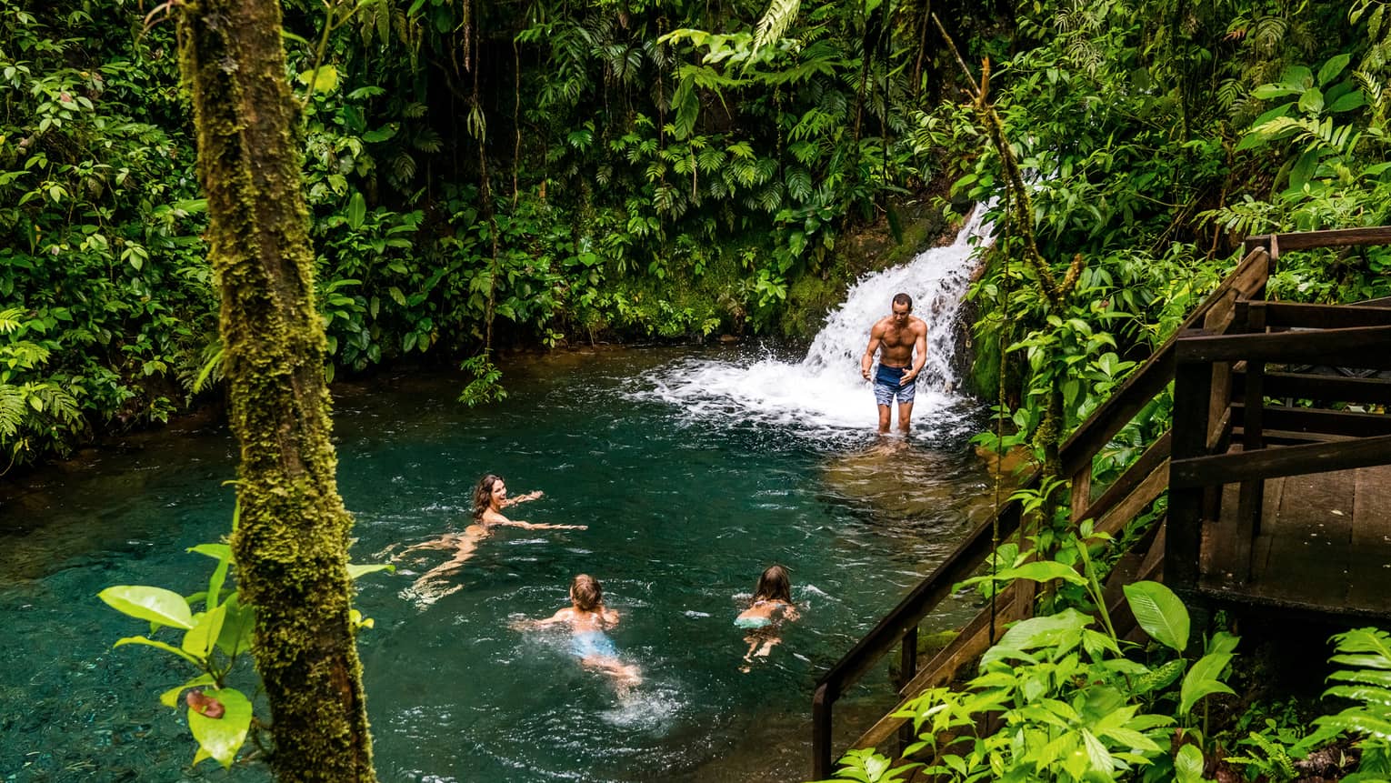 Two adults and two kids frolic at the foot of a waterfall surrounded by lush jungle greenery