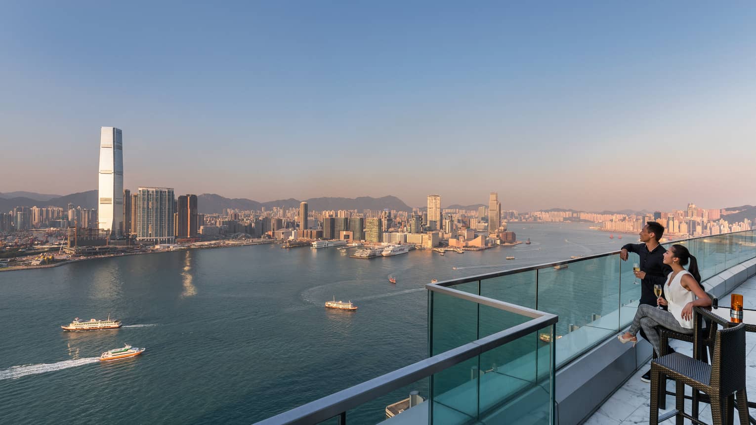 Couple on glass-walled terrace holding glasses of white wine, smiling and looking out over Hong Kong's Victoria Harbour at sunset