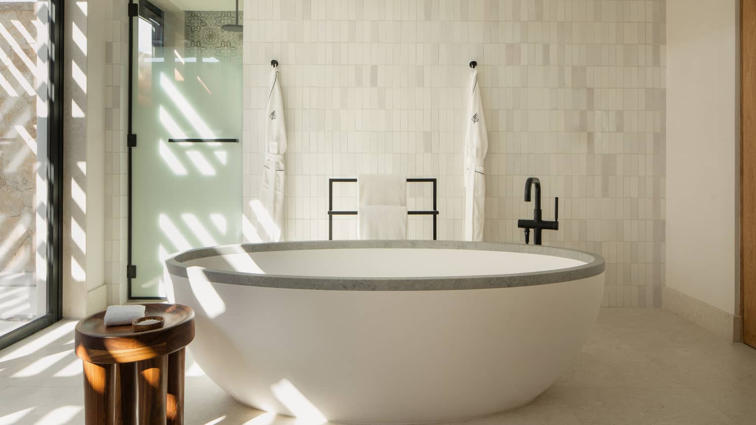 Standalone oval tub in luxury hotel bathroom, and frosted glass door leading to a separate shower. Two bathrobes hang on tiled wall's hooks.