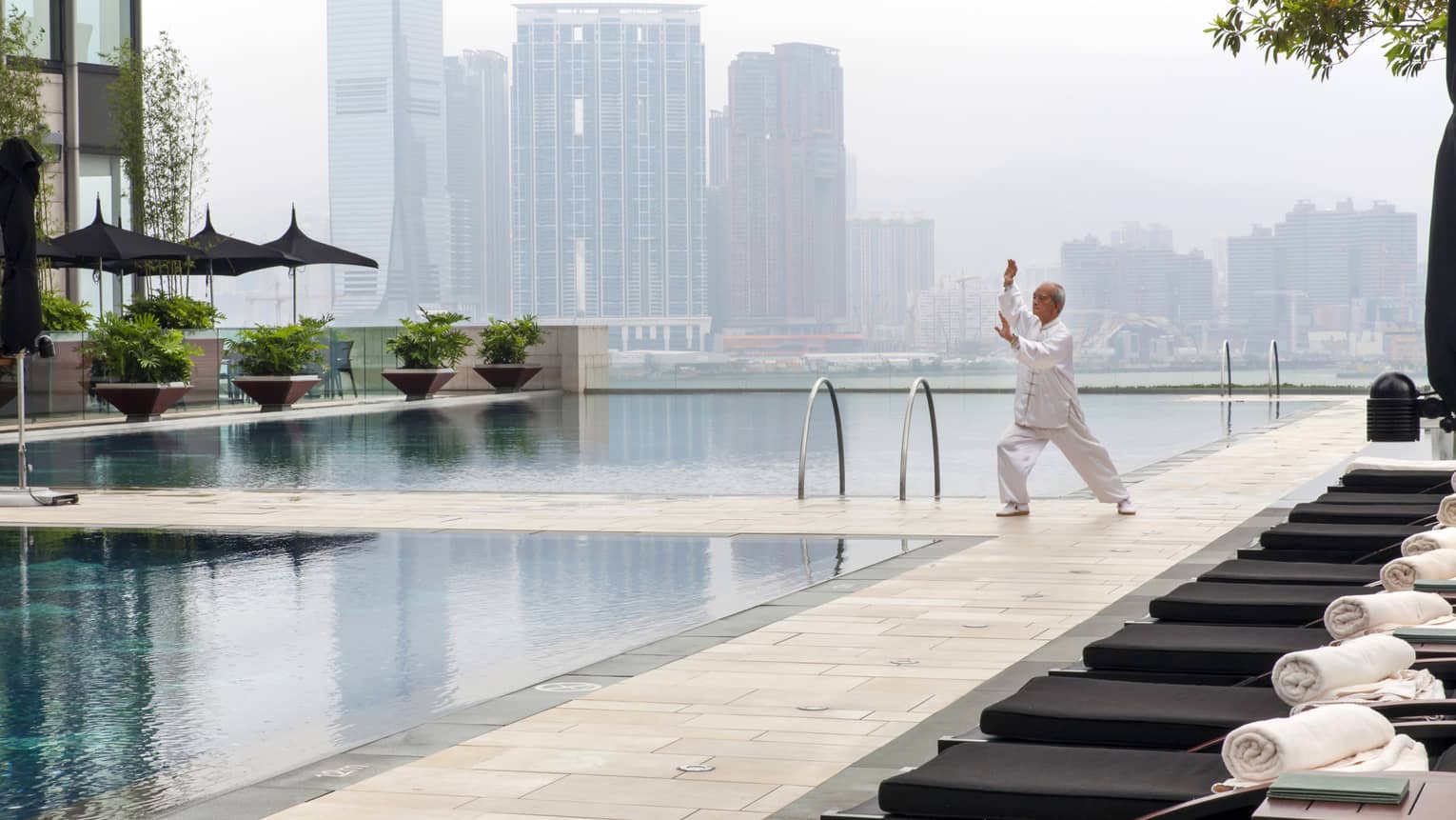 A person practices Tai Chi by a rooftop pool surrounded by greenery, with city skyscrapers in the background. The pool area has black loungers with white towels.