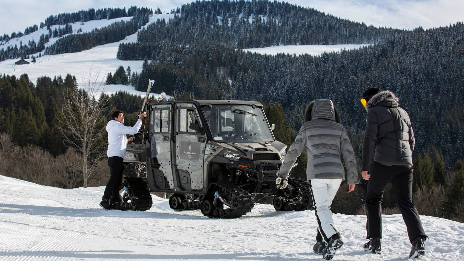 Two people in snowsuits walk toward ski concierge at large snowmobile