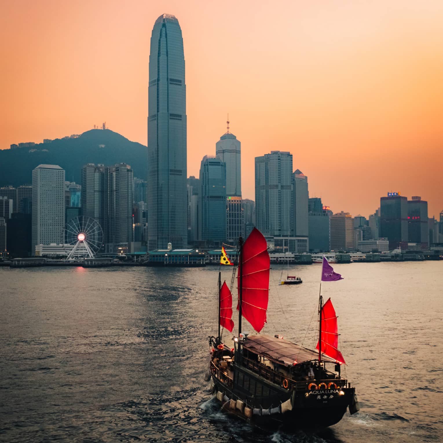 Boat with red sails on harbour in Hong Kong at sunset with city buildings at the shore