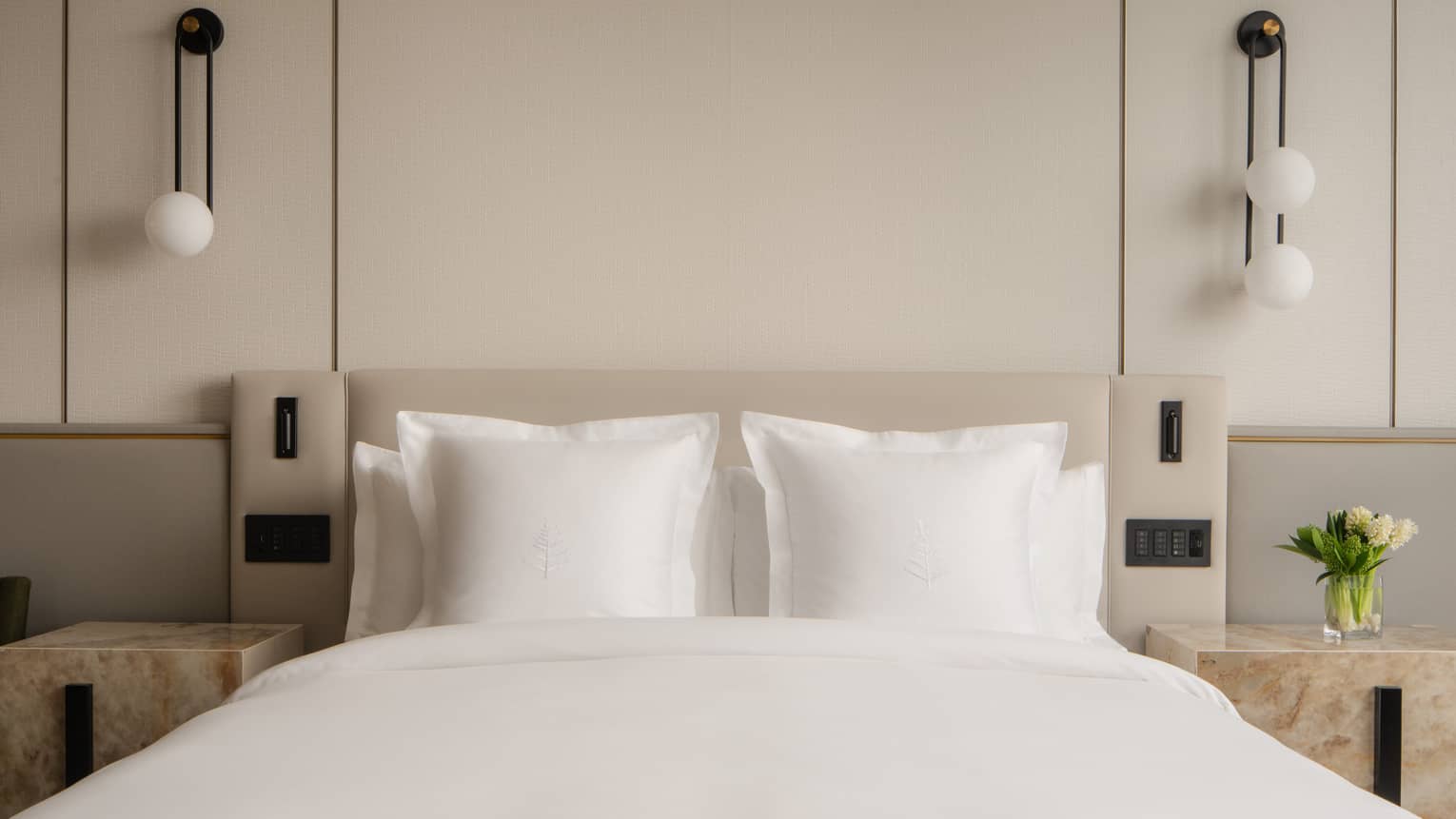 Close-up of a hotel room bed with white pillows, nightstands, bedside controls, a vase with flowers and pendant lights 
