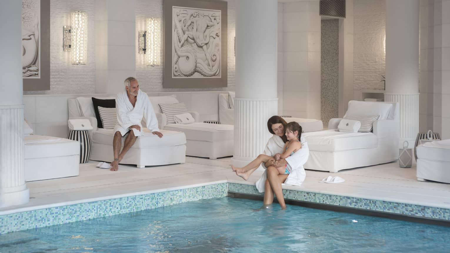 A child sits on her mother’s lap at the edge of an indoor pool as another guest smiles at them from a plush lounge chair.