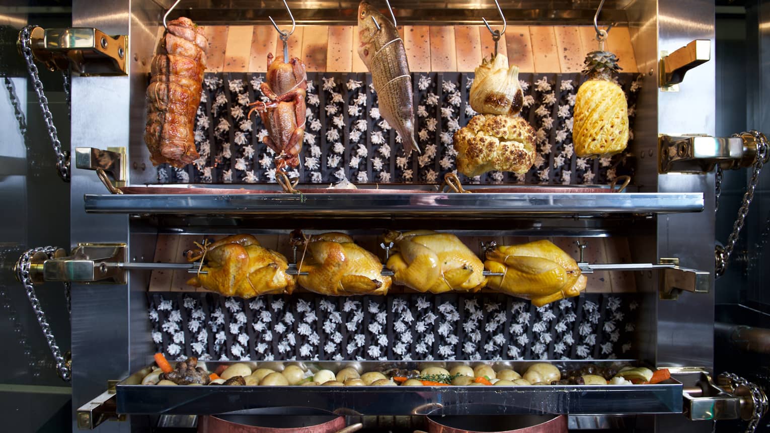 Whole chickens on rotisserie under whole fish, cauliflower hanging from hooks