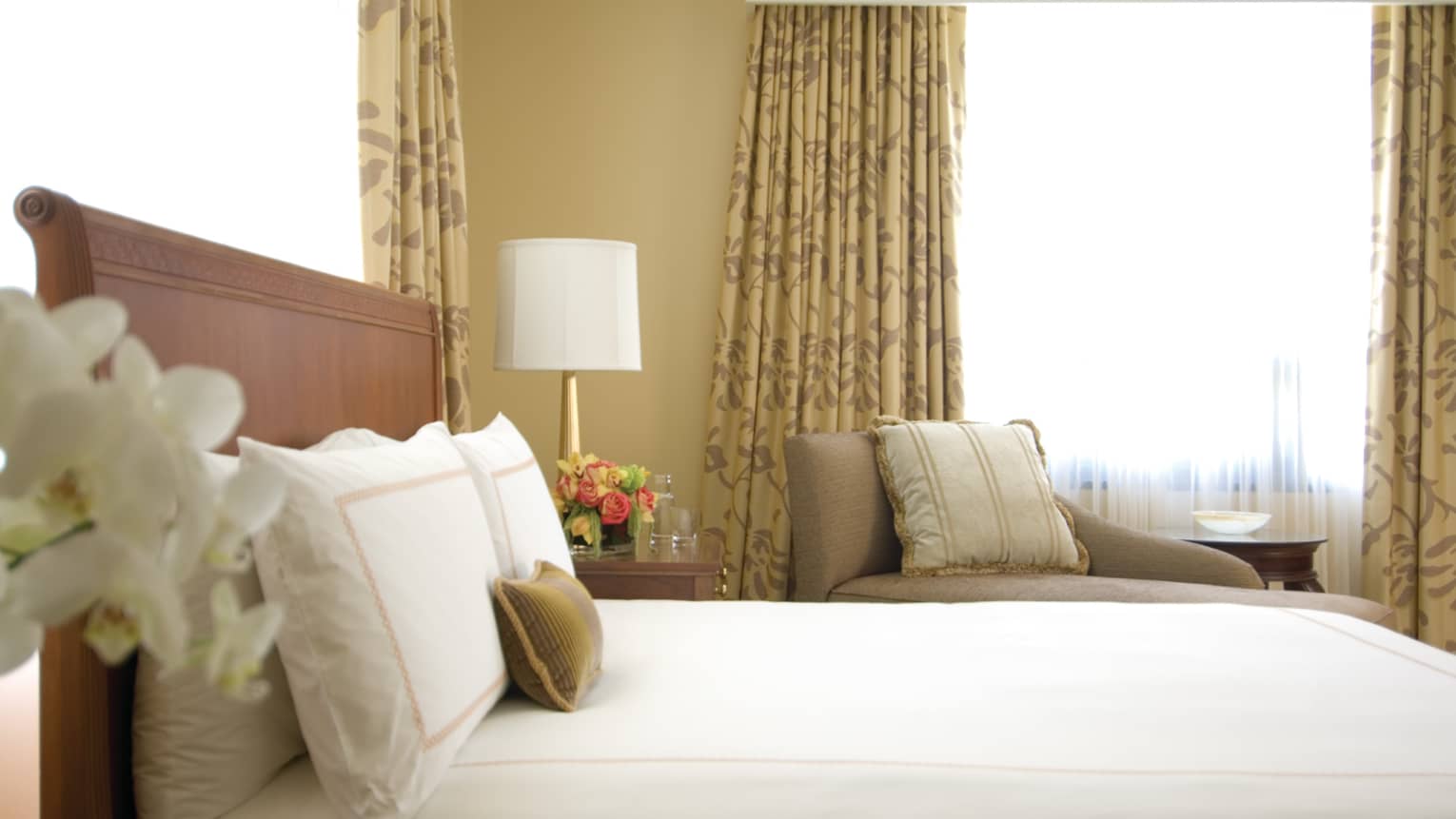 Deluxe City-View Room white orchids, side of bed, pillows against headboard, armchair, window in background