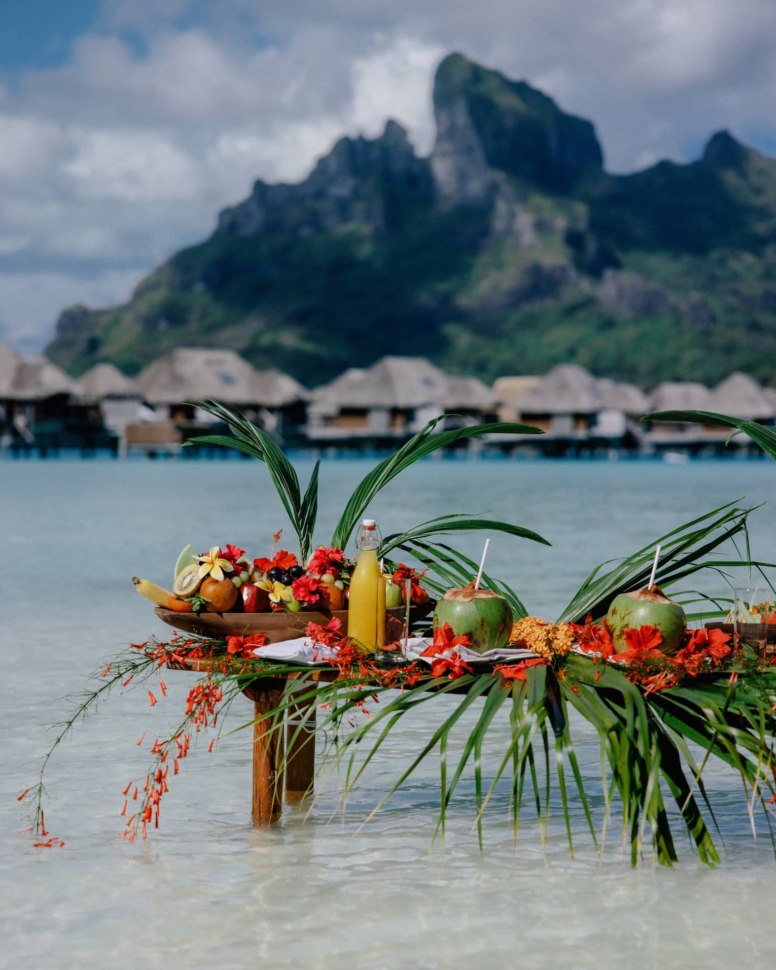 Small wooden table with legs submerged in clear water, topped with assorted fruit and coconuts with straws, mountain and overwater pavilions in background