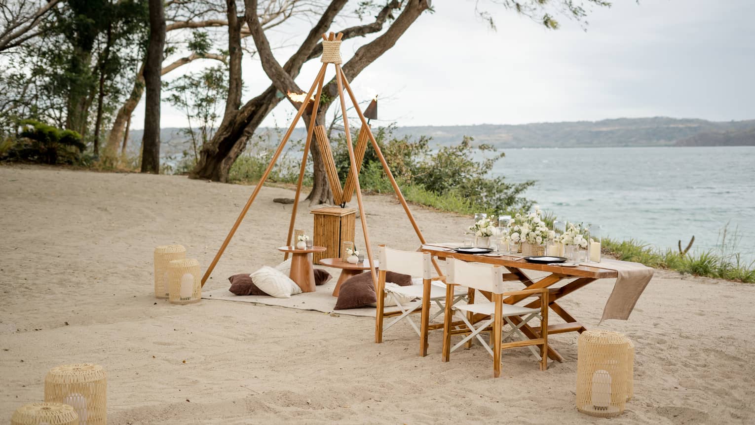 Beach dining set up with table, chairs, blanket, pillows, lanterns and tiki torches