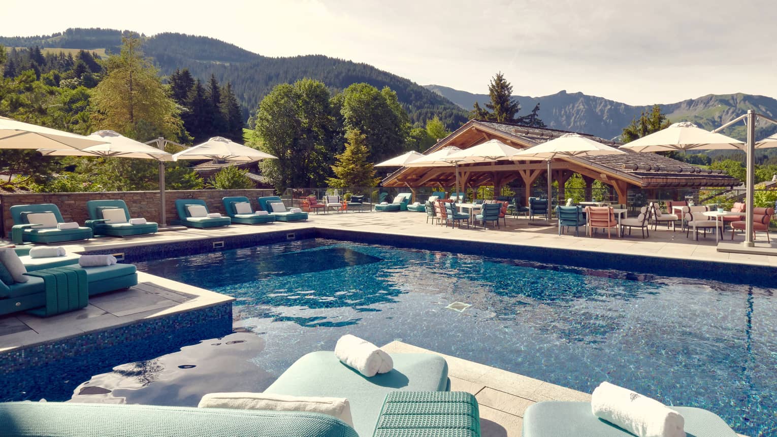Outdoor pool with aqua blue chaises longues and beige umbrellas on surrounding deck and mountains in background