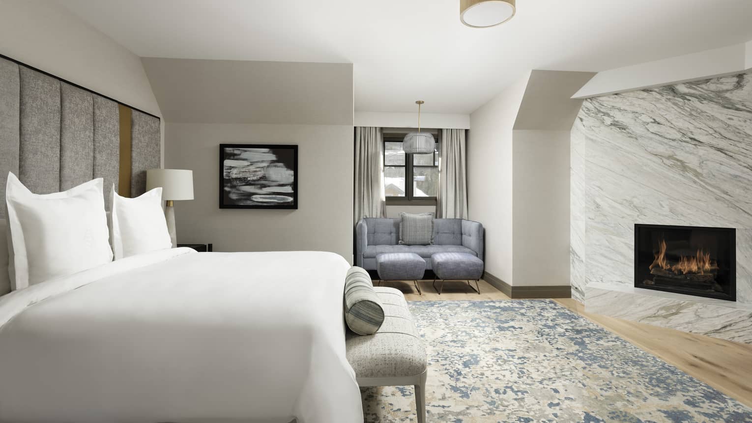 Suite bedroom with King bed and white. linens, built-in fireplace in marble wall, light blue patterned area rug, reading nook with blue loveseat
