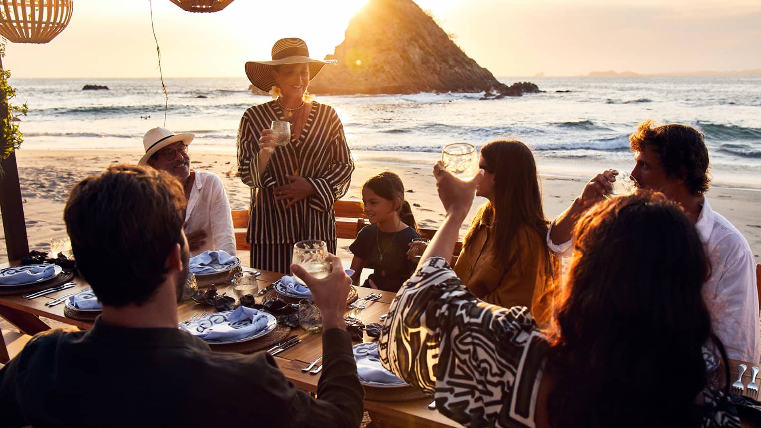 At a set table on the beach, the sun setting over the water behind them, a group of guests raise their glasses in a toast.