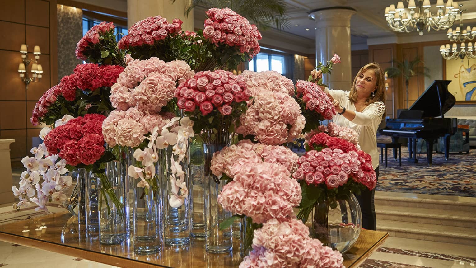 Woman arranges flowers on lobby table filled with vases, pink roses and floral arrangements 