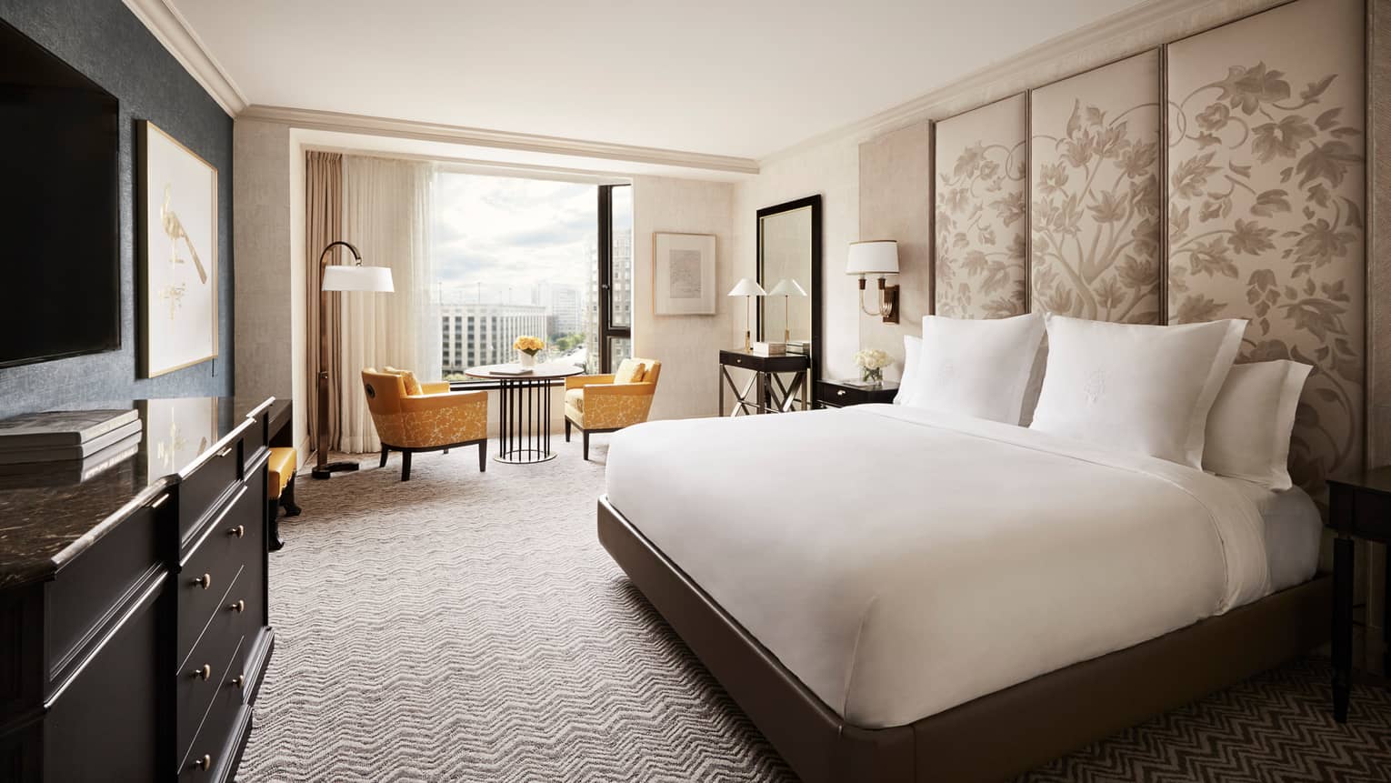 Deluxe City-View Room white bed with tall floral fabric headboard, black dresser, yellow velvet dining chairs by window