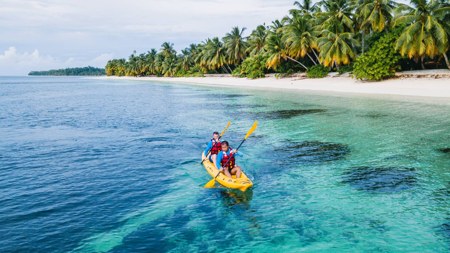 Two men wearing life jackets in yellow kayak paddle in ocean with palm trees in background