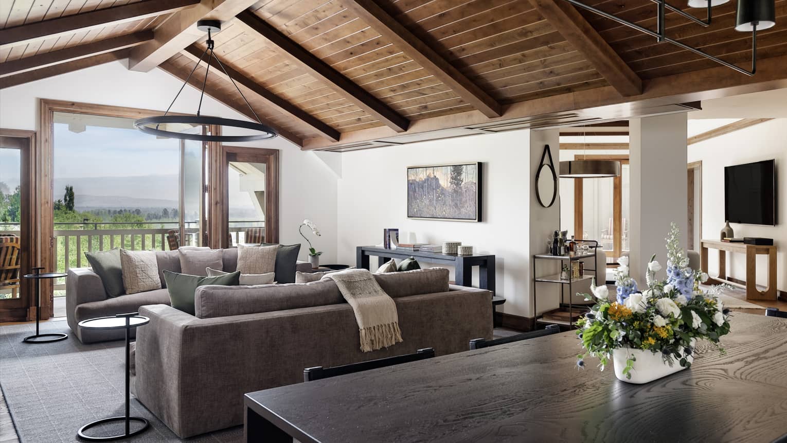 Living and dining area with vaulted wood ceiling, two grey sofas, dining table with a floral centrepiece, and mountain views