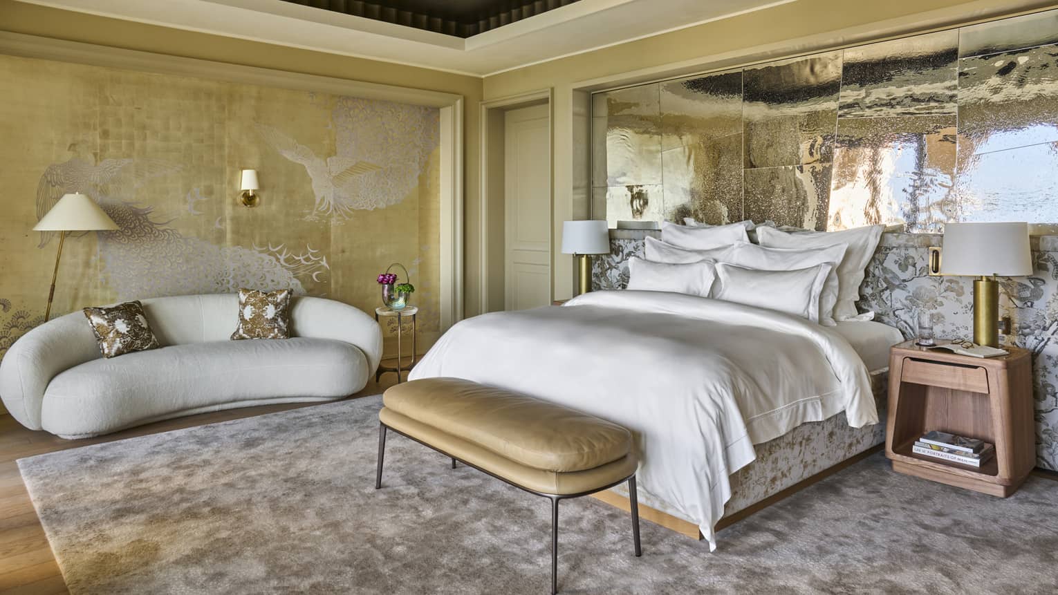 Large hotel suite bedroom featuring king bed with white linens, gold upholstered bench, side tables and lamps, white curved sofa and a silver mirrored accent wall,Large hotel suite bedroom featuring king bed with white linens, gold upholstered bench, side tables and lamps, white curved sofa and a silver mirrored accent wall