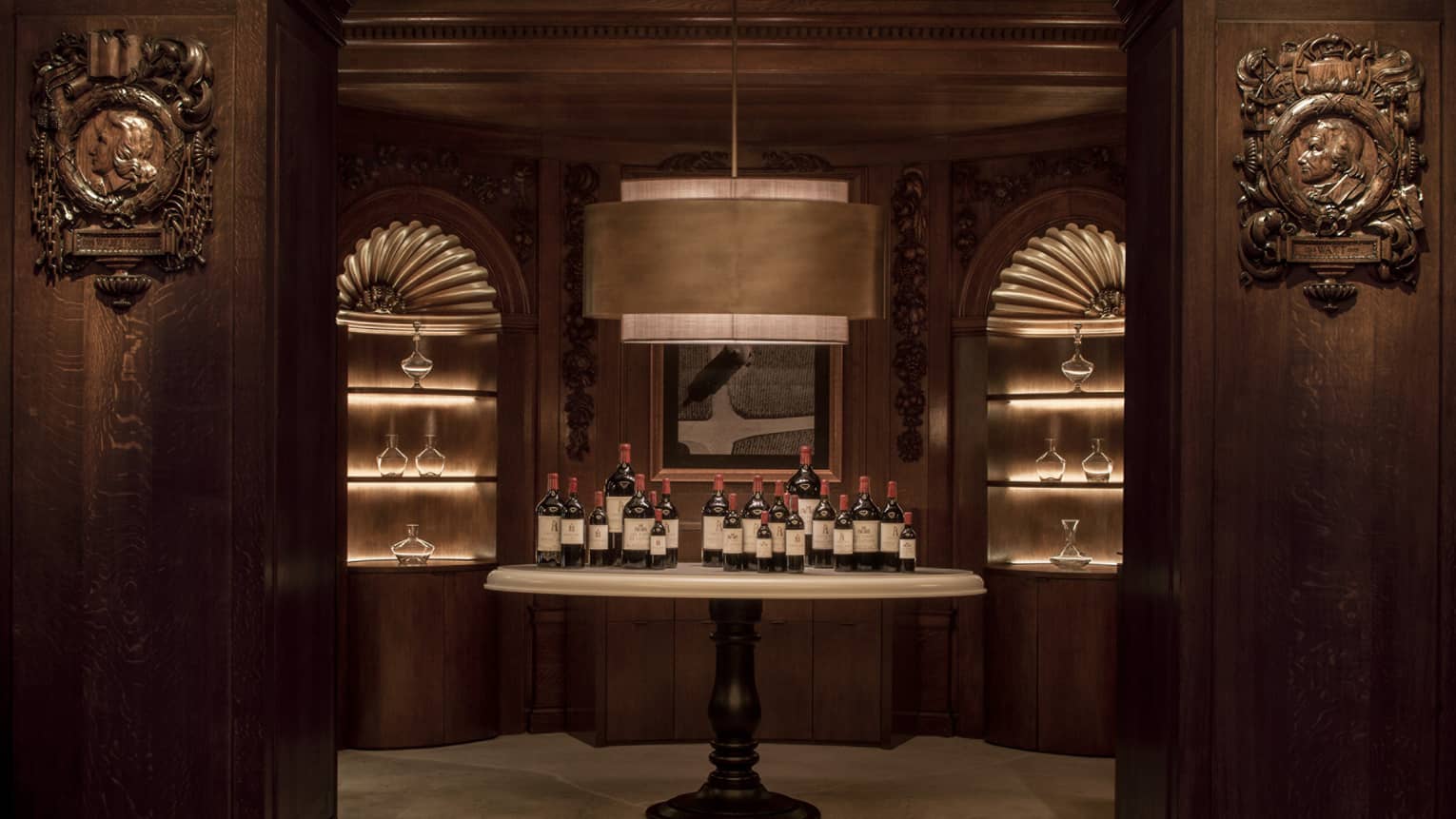 In a dark-panelled room with a large, stylish pendant lamp, a pedestal table holds twenty bottles of wine in varying sizes.