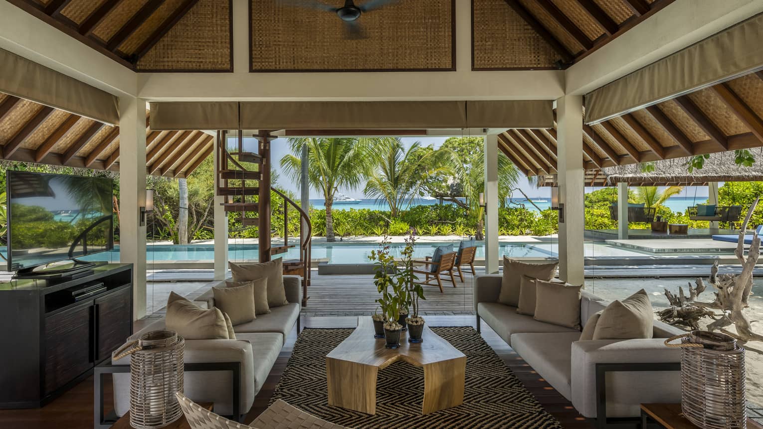 A covered outdoor living room with a television, plush couches and pool access