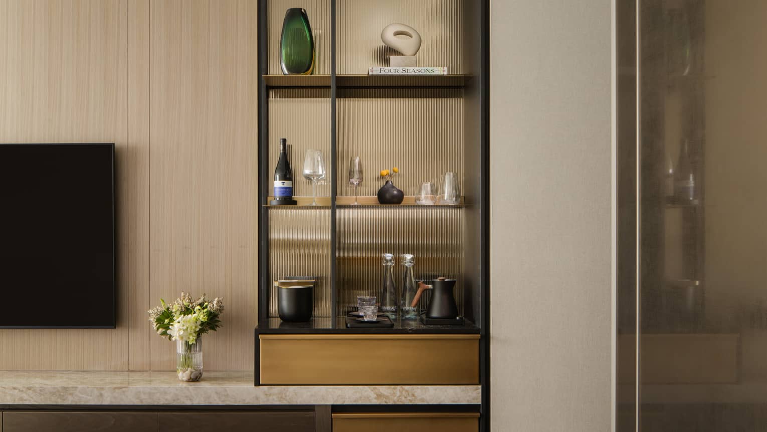 Stylish minibar with glass shelves displaying wine, glasses, and decorative items, next to a TV and vase with fresh flowers
