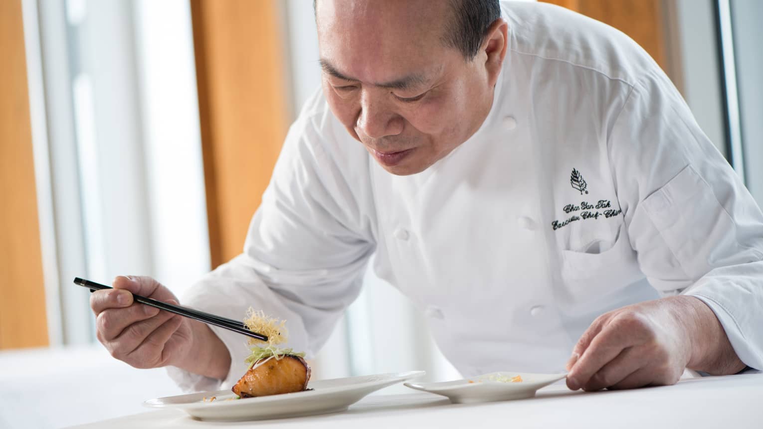 Chef in white uniform leans over grilled salmon dish on plate, adds garnish with chopsticks