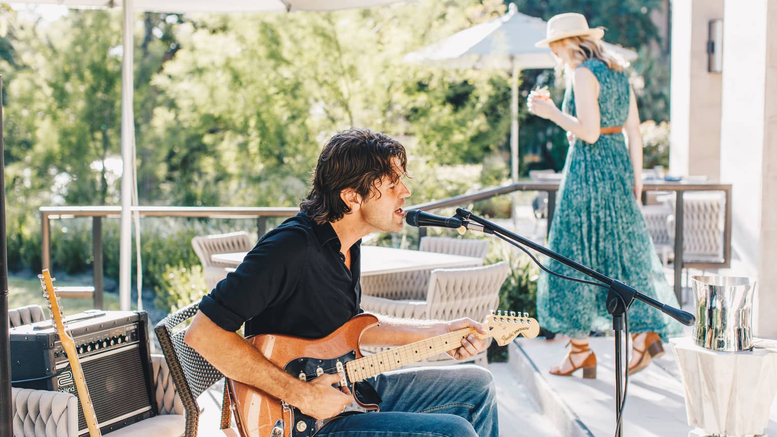 A man sitting and playing guitar while a woman in a green dress walks behind him.
