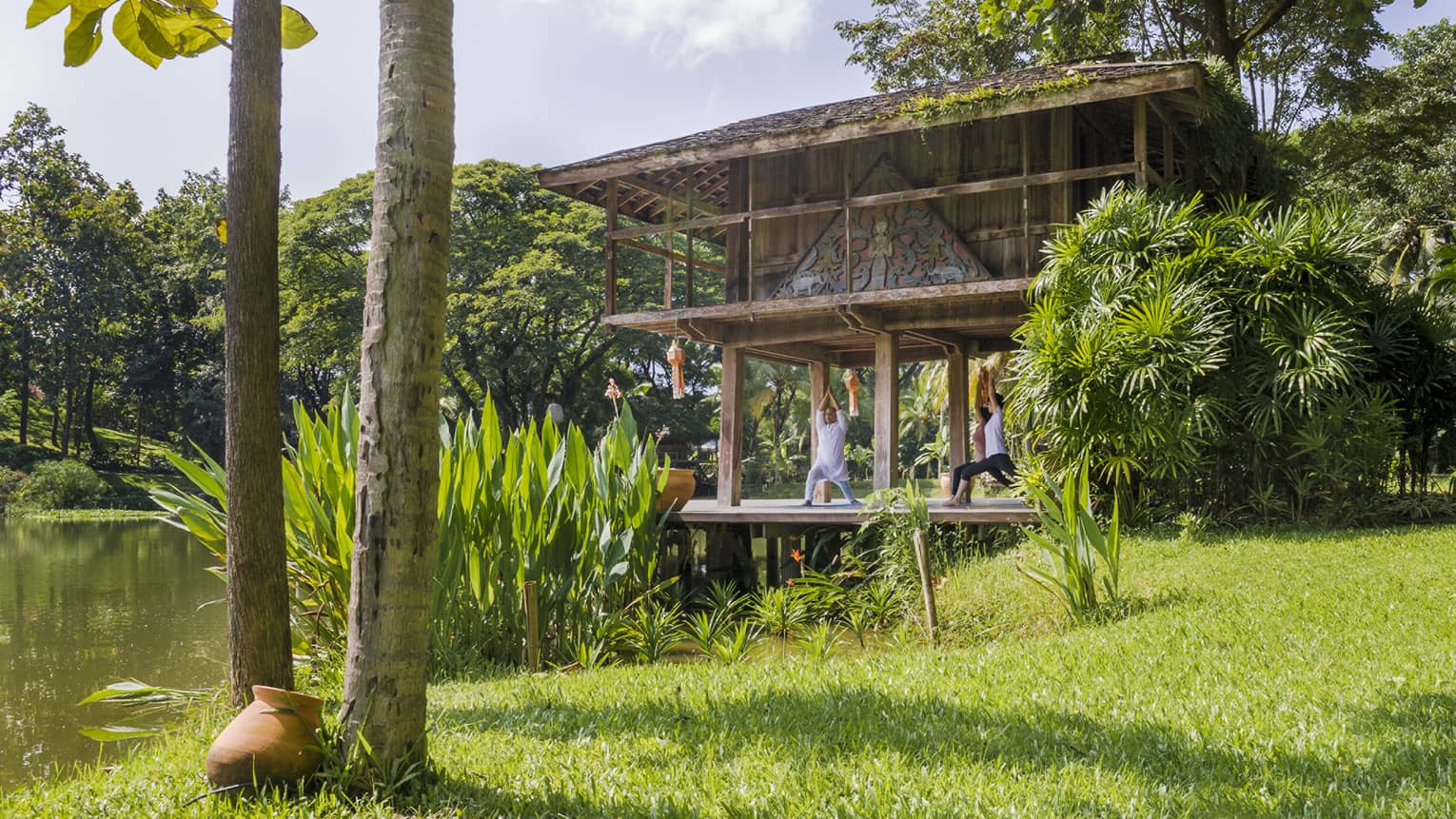 Yogi and two guests kneel with arms raised in yoga posed on pavilion deck near pond