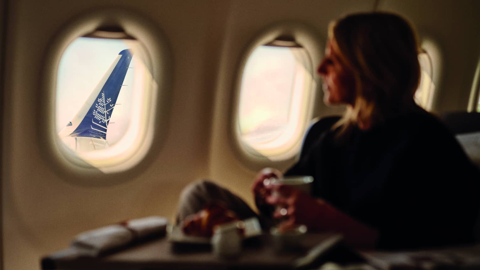 Person with shoulder-length blonde hair wearing a dark sweater and pants holds a beverage as they gaze out a plane window overlooking the wing
