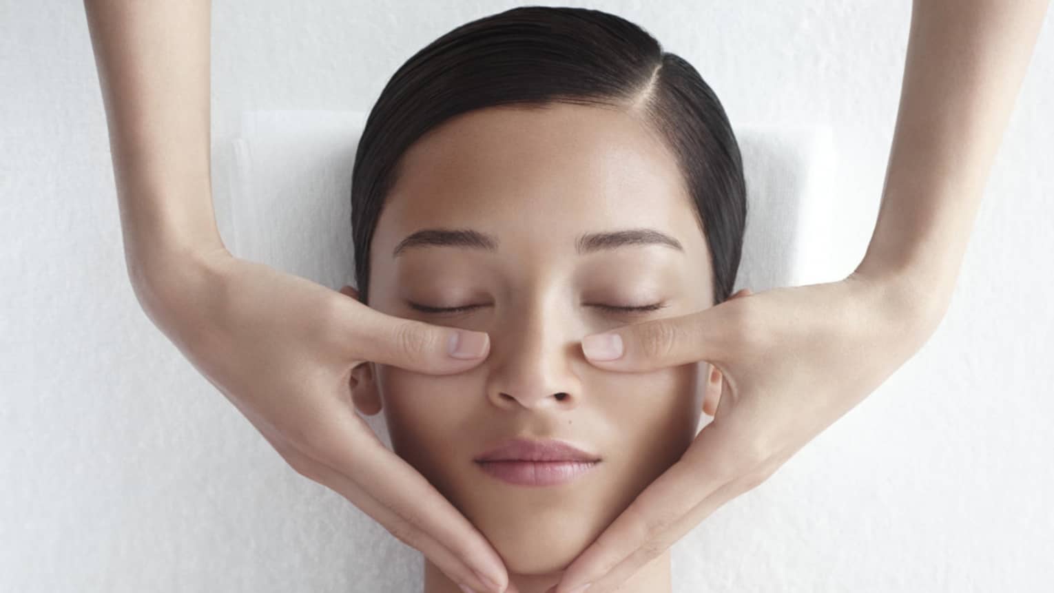 Woman in white bathrobe lies on massage table with eyes closed, masseuse rests hands on her cheeks