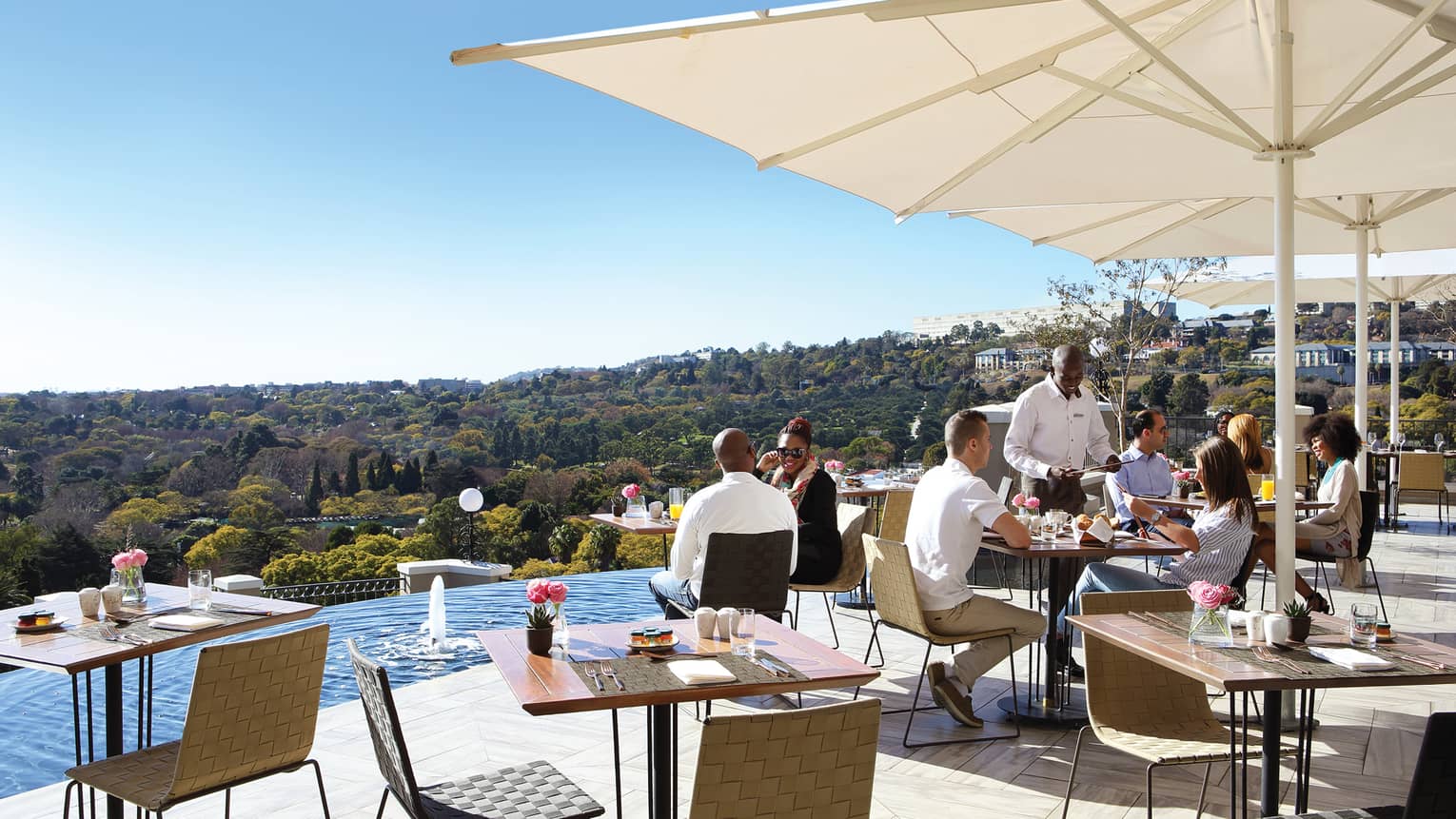 People socialize around patio dining tables on sunny deck near swimming pool