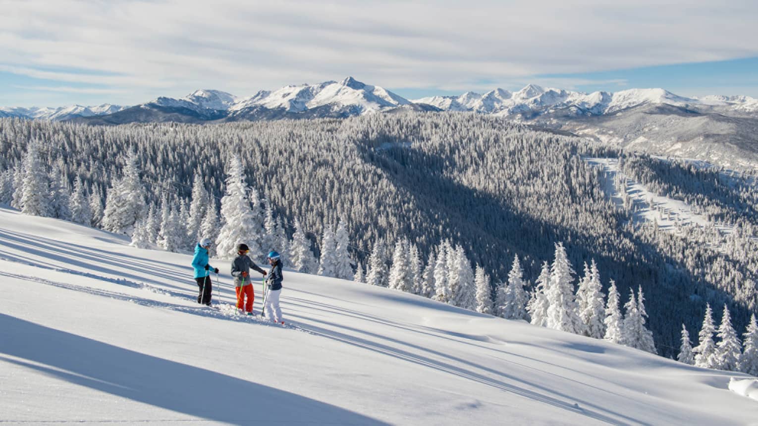 Three guests on skis pause on the ski hill, snow capped trees and mountains in distance 