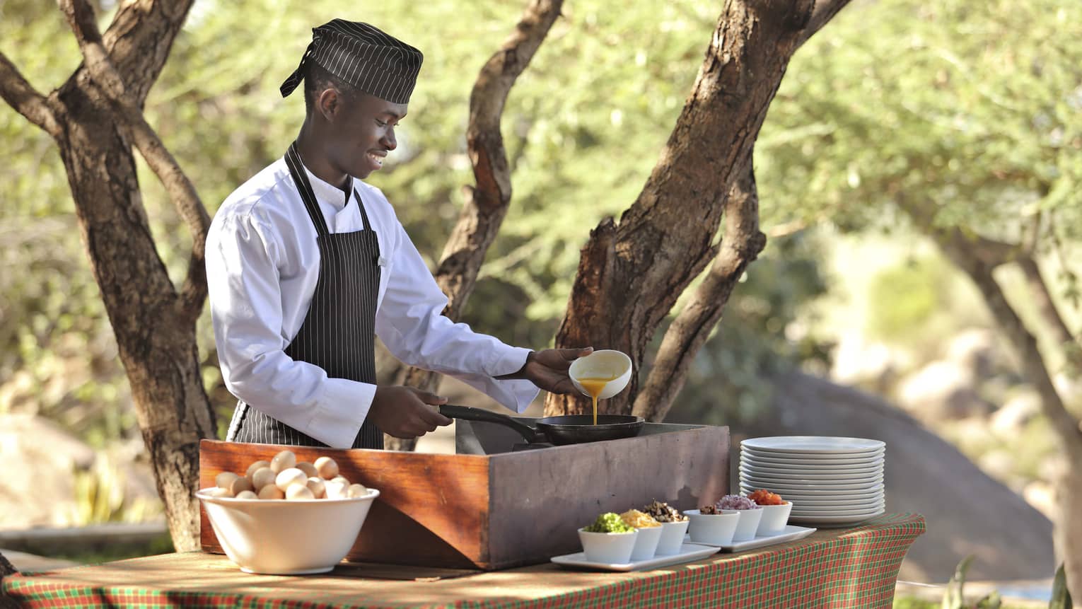 Smiling chef prepares eggs at outdoor breakfast station beside tree 
