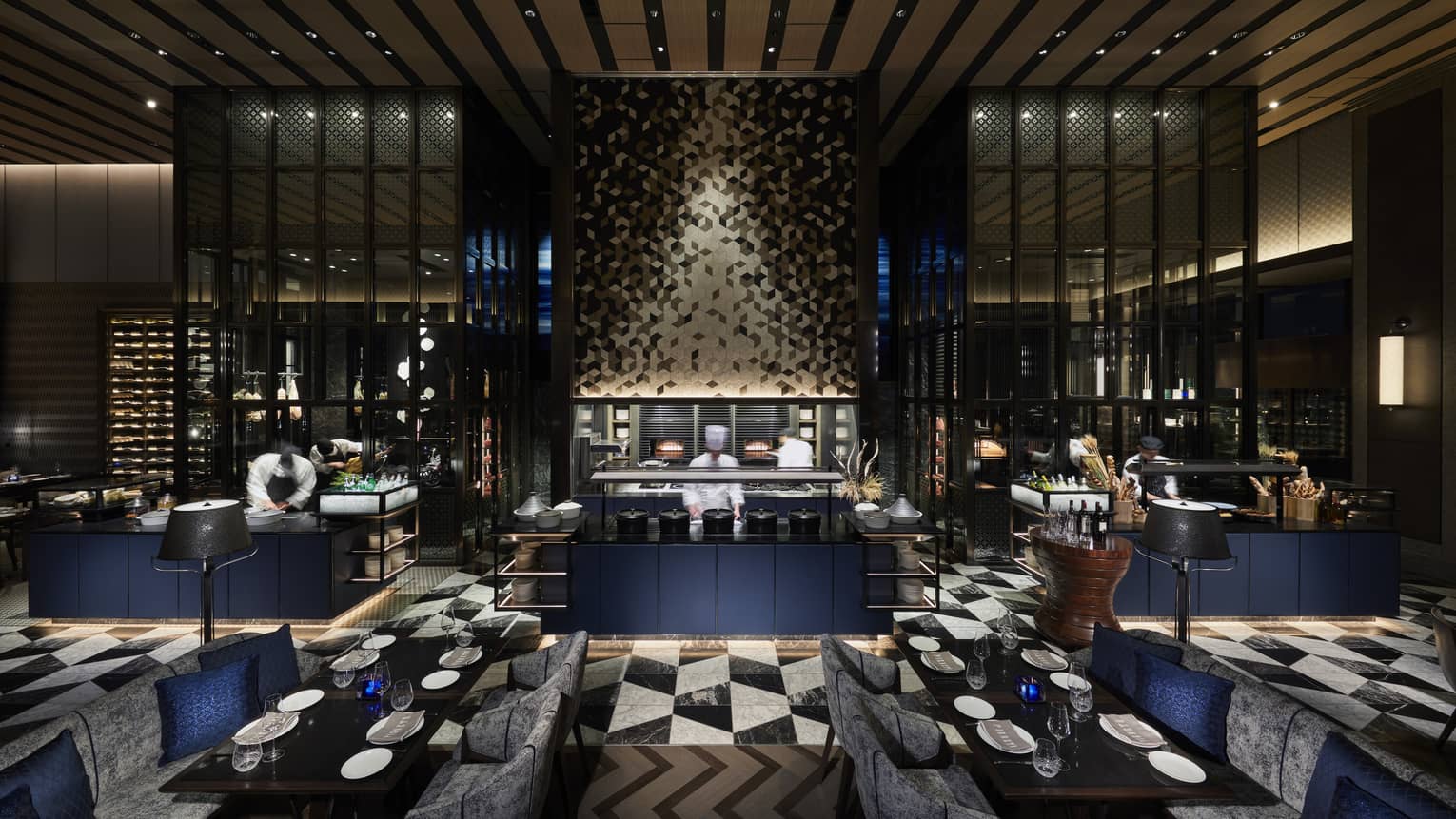 Chefs work diligently in open-air kitchen, tables set for six in the foreground, grey and black colour scheme 