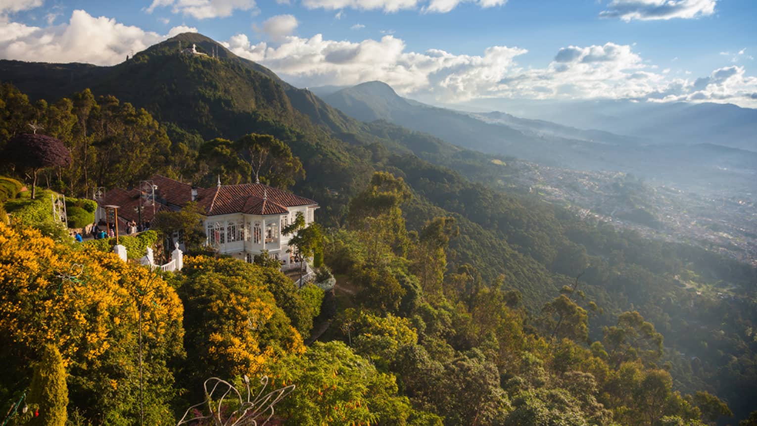 Aerial view of colonial-style white building tucked high on lush green mountain that stretches to horizon, blue sky