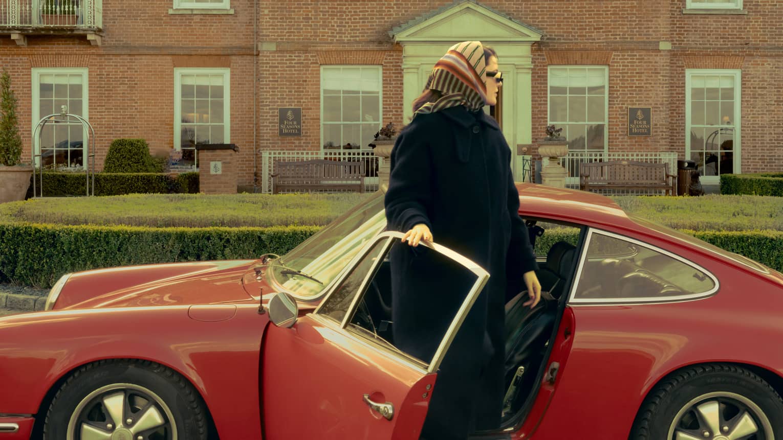 Person in black coat and head scarf getting out of a red vintage sports car on driveway in front of red brick building