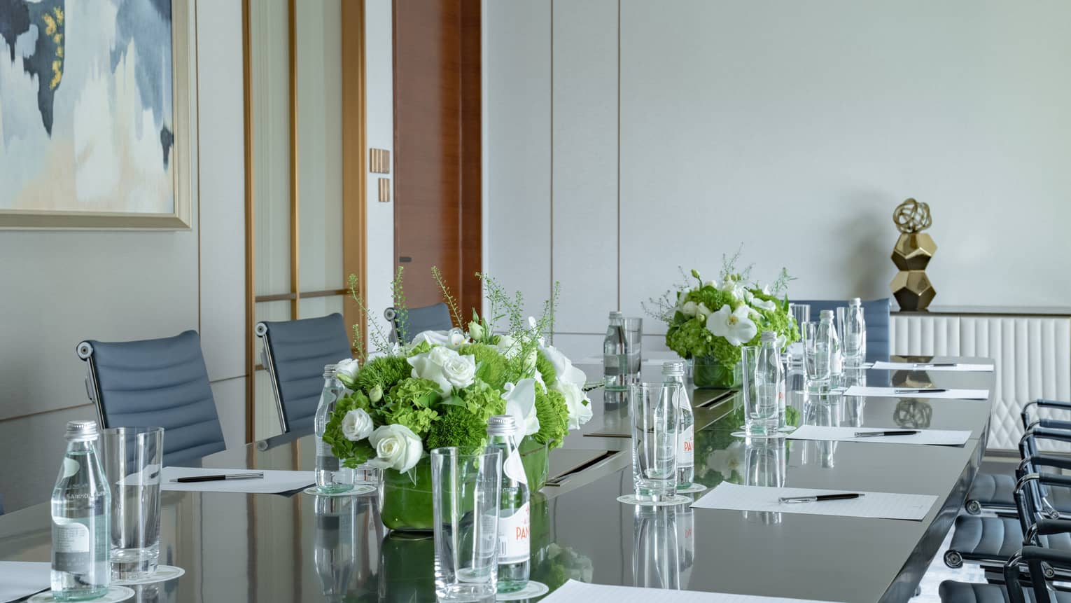 Meeting room with long boardroom table set with papers and pens, floral arrangements, bottles of water